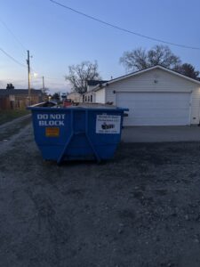 A blue dumpster from Dump N Dash Dumpsters LLC, marked "DO NOT BLOCK" and containing debris, placed in a residential area in Valley, NE.