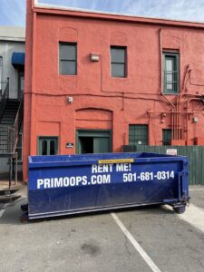 A blue dumpster from Primo Operations, available for rental, parked in front of a building in North Little Rock, AR.