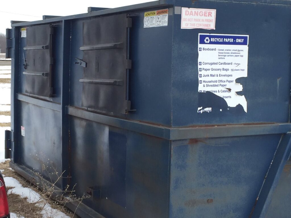A blue recycling dumpster with a 'RECYCLE PAPER - ONLY' sign from NID Dumpster Rental in Nashua, IA.
