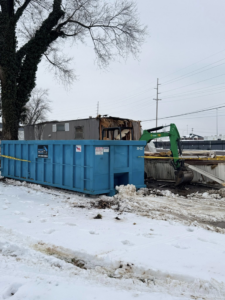 A blue roll-off dumpster from Ryan's Roll-Off Dumpsters at a snowy demolition site with an excavator in Owensboro, KY.