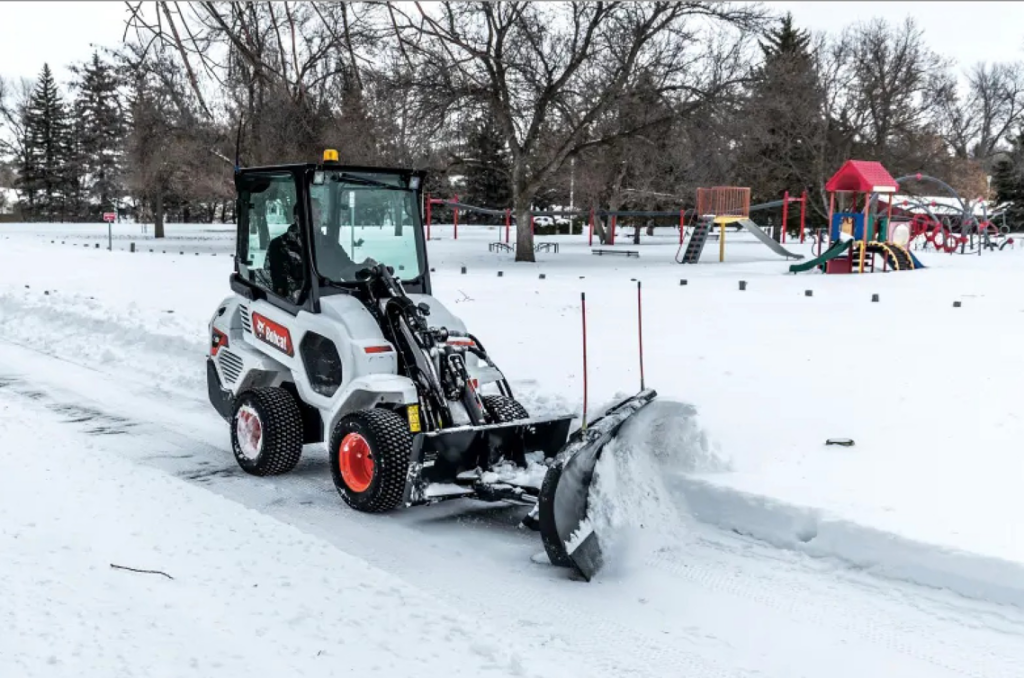 A Bobcat compact wheel loader with a snowplow attachment clearing snow in a park, from Handyman Rental in Portland, ME.