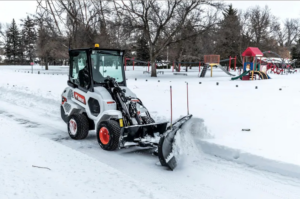 A Bobcat compact wheel loader with a snowplow attachment clearing snow in a park, from Handyman Rental in Portland, ME.