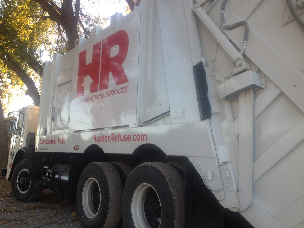 A branded grey trash can with a red lid from Husker Refuse Services in Lincoln, NE, for waste collection.