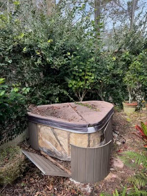 A broken hot tub partially hidden in a garden, awaiting removal by Route Junk in Orlando, FL.