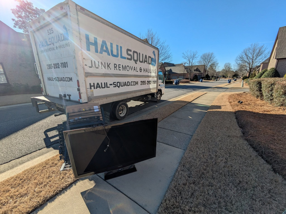 A broken flat-screen TV placed on the curb next to a Haul Squad truck for removal in Helena, AL.