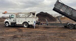 A bucket truck and a large dump trailer with a pile of wood chips in the background, used by Happy Lemon Tree Service in Belton, MO.