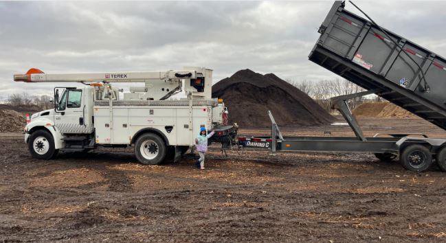 A bucket truck and a large dump trailer with a pile of wood chips in the background, used by Happy Lemon Tree Service in Belton, MO.