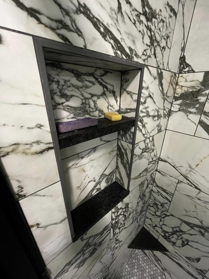 A stylish built-in shower niche with black shelves against elegant marble-look tiles by Simko Tile & Bath in Macon, GA.