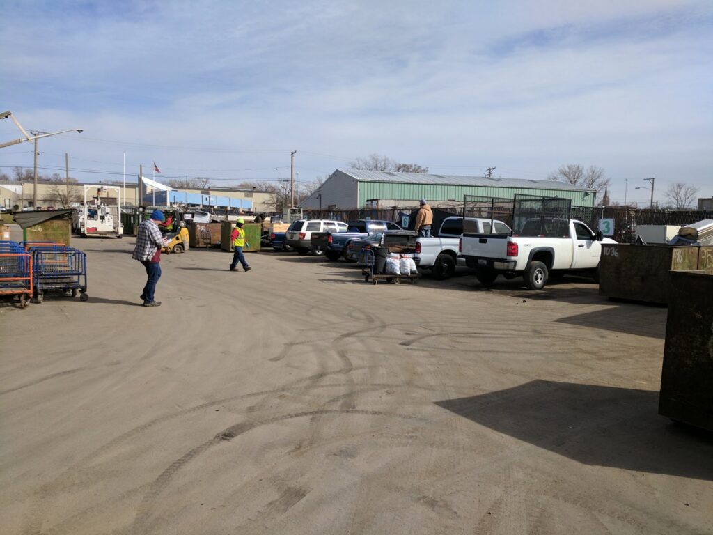 A busy outdoor recycling yard with multiple trucks, dumpsters, and workers at Get Green Recycling Co. in Aurora, IL.