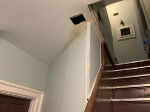 A ceiling with a small square hole and a visible water stain on the adjacent wall near a staircase, showing a repair need by PG Painting and Contracting Services in Cincinnati, OH.