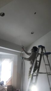 A handyman wearing a respirator, spray painting a ceiling while on a ladder, by MDJ Handyman in Salt Lake City, UT.