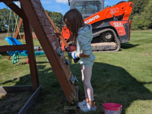 A child using a power drill to work on a wooden playset climbing wall, a project by Woods Builders in Grand Rapids, MI.