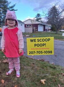 A child in a costume standing next to a 'We Scoop Poop!' sign for The Doo Doo Dudes in Lewiston, ME.