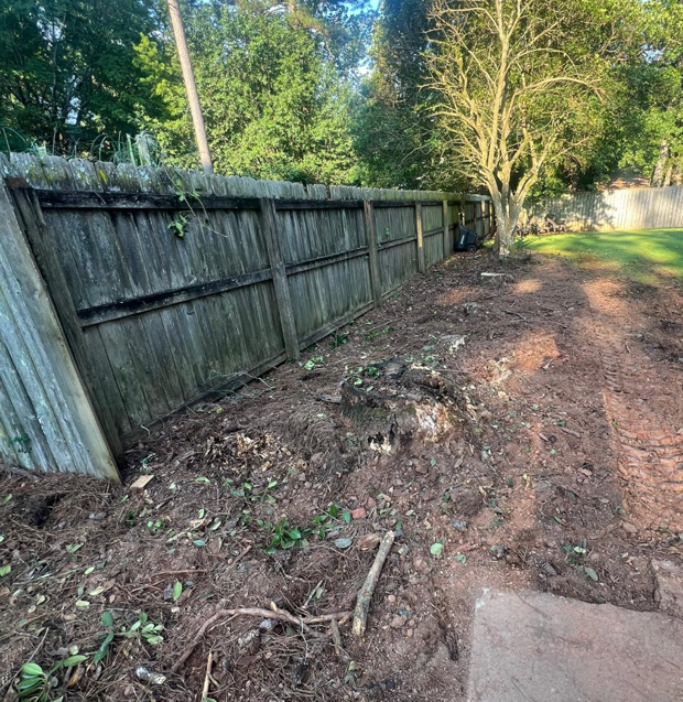 A cleared area next to a wooden fence, showing the ground after a stump removal or tree clearing service by Manley's Stump Grinding in Opelika, AL.
