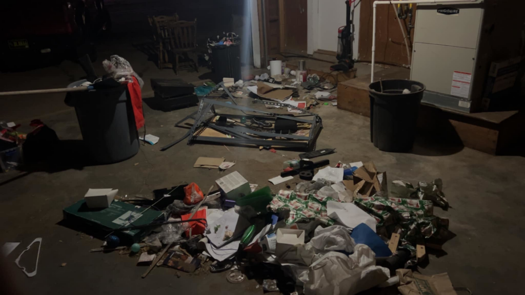 A dimly lit garage or basement floor covered in trash, debris, and various unwanted items before cleanup by 575 Junk Removal in Clovis, NM.