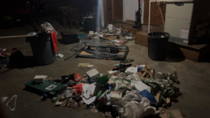 A dimly lit garage or basement floor covered in trash, debris, and various unwanted items before cleanup by 575 Junk Removal in Clovis, NM.