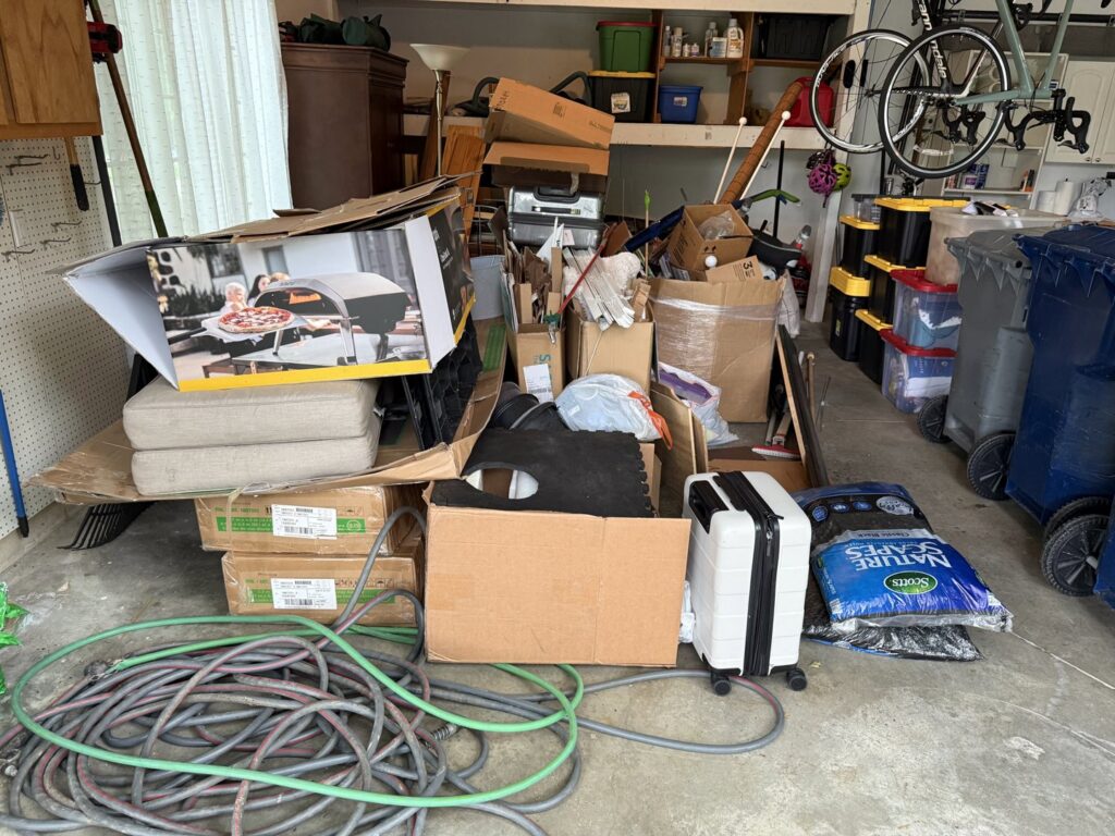 A cluttered garage filled with boxes, hoses, and various items, indicating a junk removal service by Indy Trash Guy in Fishers, IN.