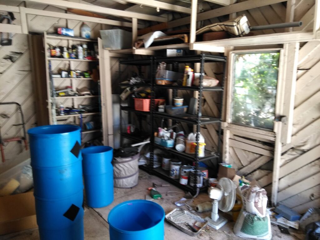 A cluttered garage filled with shelves, blue barrels, and various hazardous waste items awaiting removal by Hazawaytoday.com in El Segundo, CA