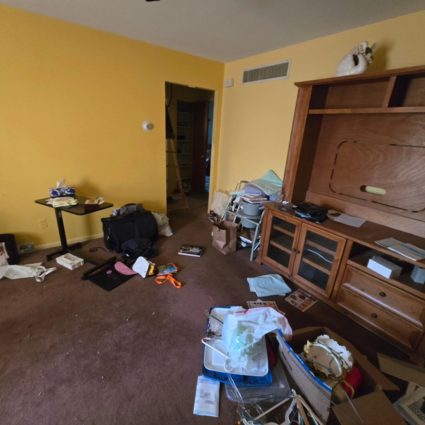 A cluttered living area with yellow walls and brown carpet, awaiting a cleanout or renovation by 1st State's Finest LLC, a handyman in Middletown, DE.