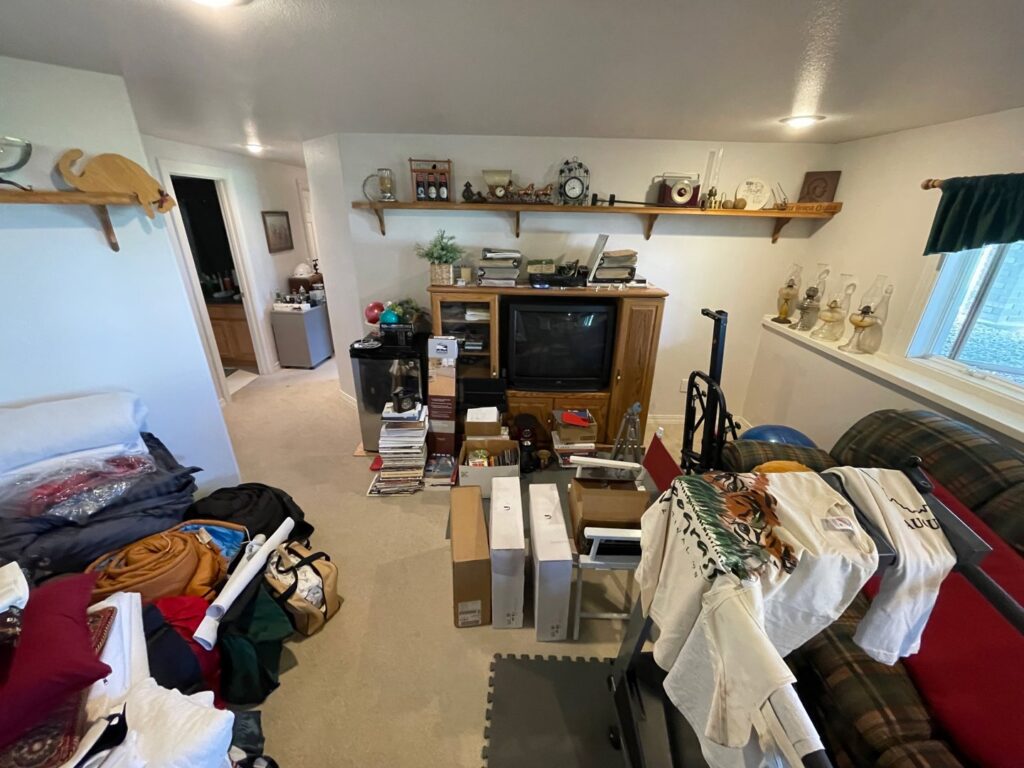 A cluttered room with furniture, boxes, and miscellaneous items, showing a 'before' scene for a junk removal cleanout by Empty It Out Junk Removal, LLC in Billings, MT.