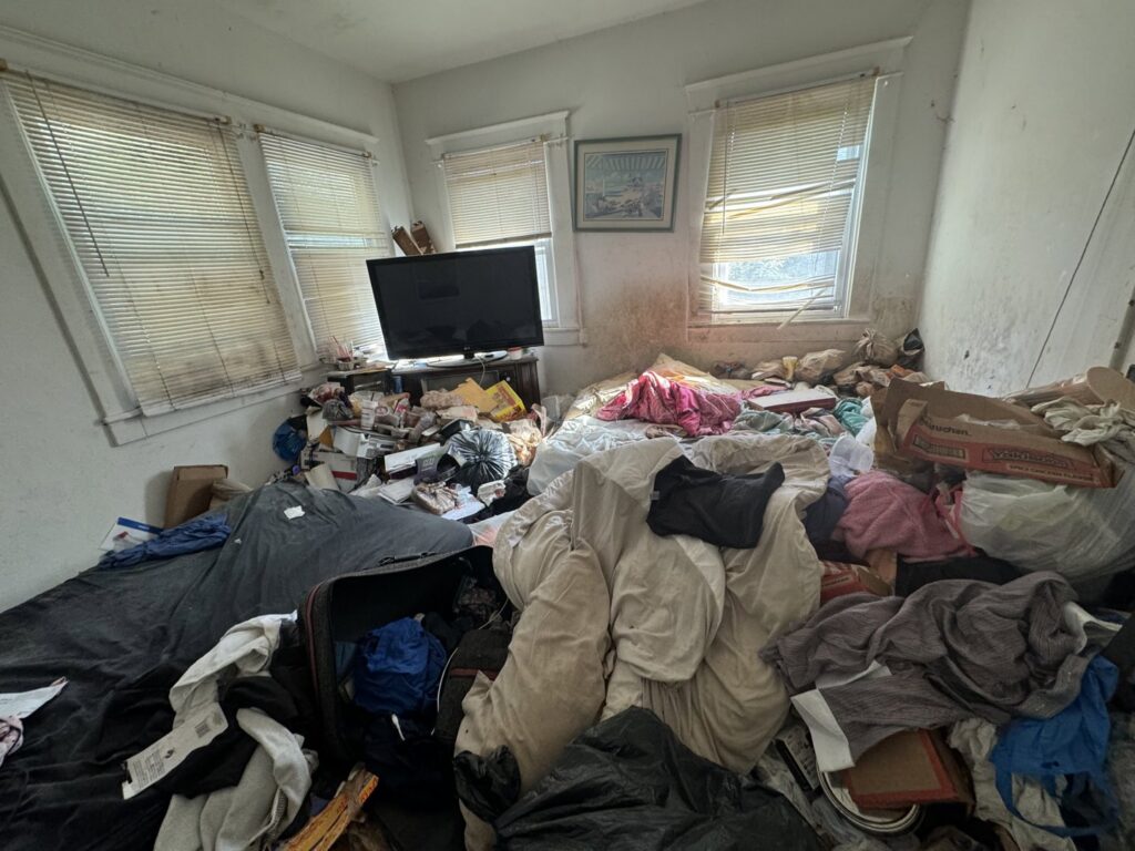 A heavily cluttered room filled with clothes, boxes, and debris, indicating a cleanout job for Music City Removal in Nashville, TN.