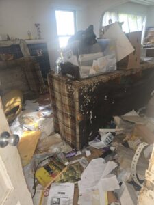 A heavily cluttered room filled with furniture, papers, and trash, indicating an estate cleanout by Titan Junk Co. in San Diego, CA