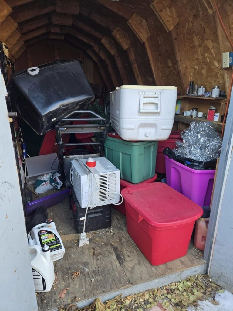 A cluttered shed interior filled with household items, boxes, and an AC unit, indicating a cleanout job for Big Sky Junk Removal LLC in Billings, MT.