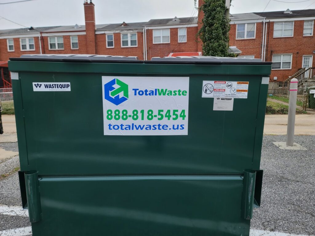 A green commercial dumpster from Total Waste located in an alleyway behind buildings in Baltimore, MD.
