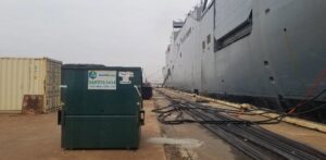 A green commercial dumpster from Total Waste positioned next to a large ship at a dock in Baltimore, MD.