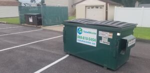 A green commercial dumpster from Total Waste in a fenced parking area with a garage in the background in Baltimore, MD.