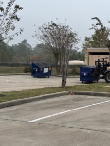 Two blue commercial dumpsters in a parking lot, with one being moved by a Waste Connections truck in Houston, TX.