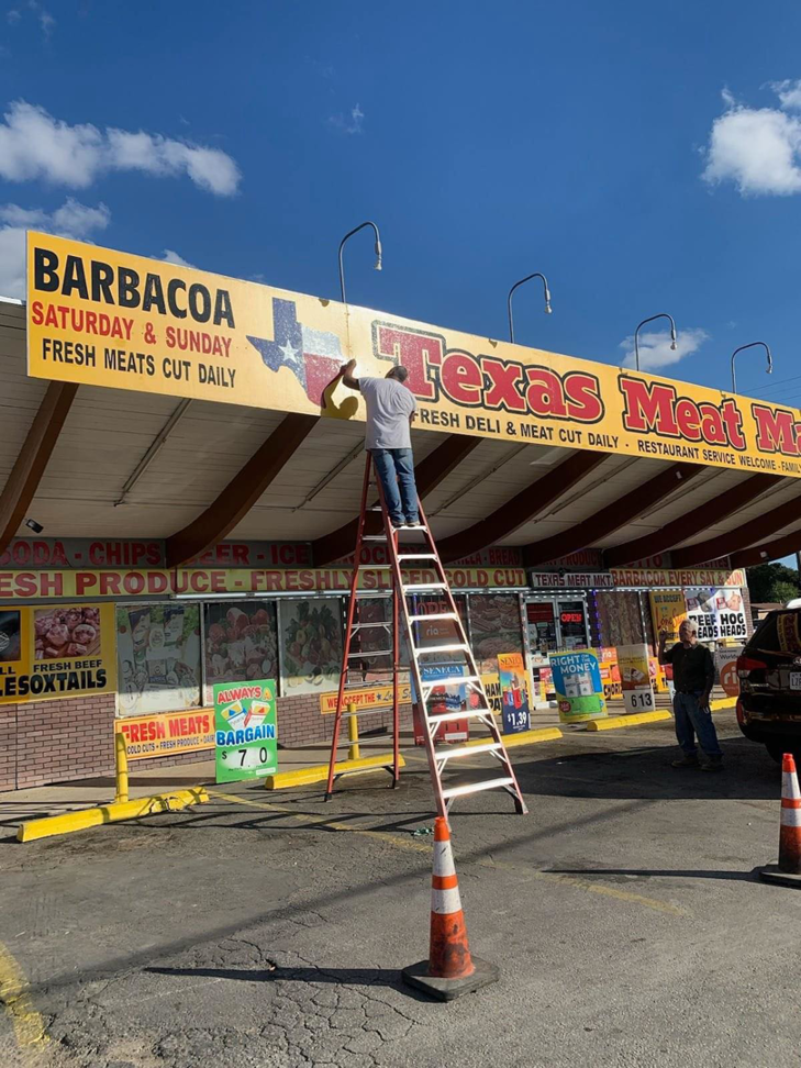 A worker on a ladder installing or repairing a commercial sign for Texas Meat Market by DeLeon Contracting LLC in San Antonio, TX.