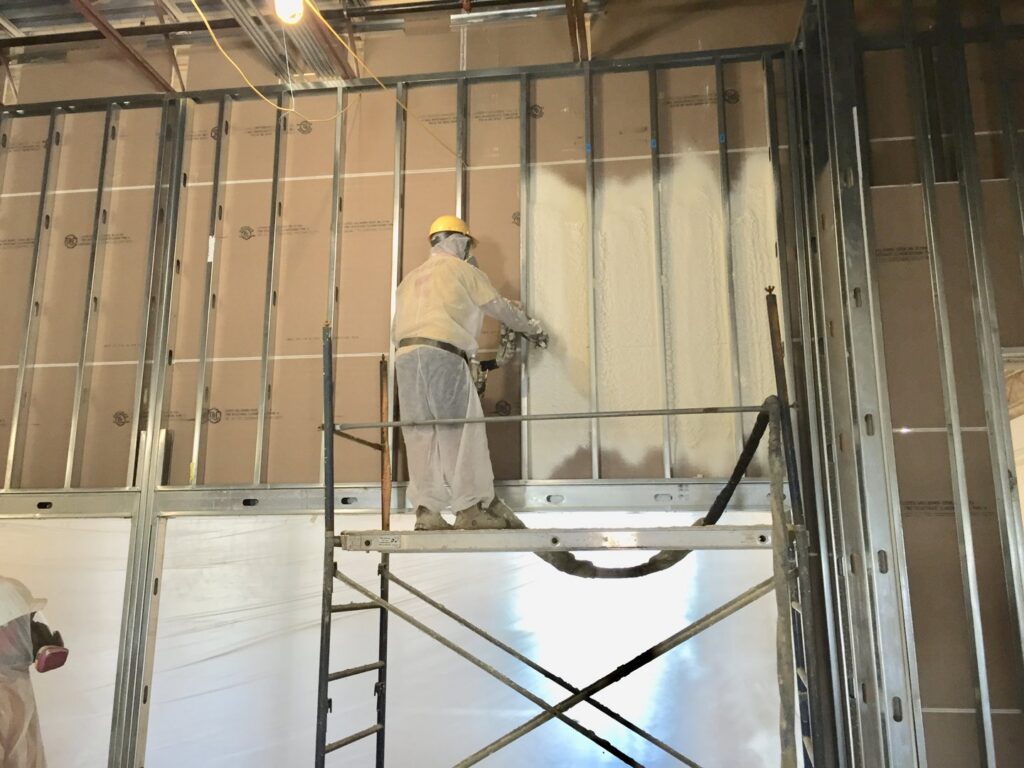 A worker on scaffolding applying spray foam insulation to a commercial building wall by Spray Foam Art-Insulation in Stamford, CT.