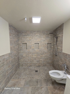A beautifully completed bathroom renovation featuring a new tiled walk-in shower and toilet by Torres Services in Anchorage, AK.
