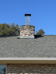 A close-up of a newly completed roof with a stone chimney and dark shingles, showcasing work by APEX Roofing in Georgetown, TX.