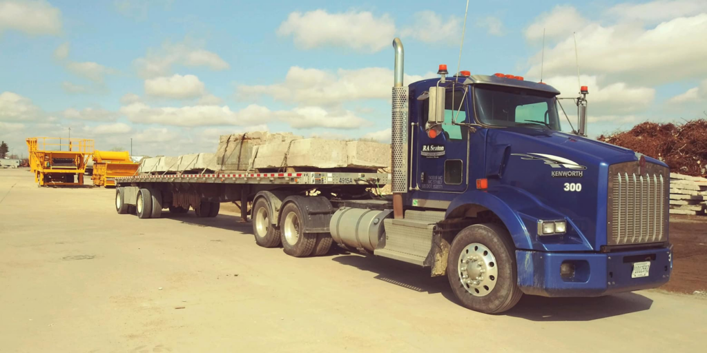 A blue R A Seaton Contractor Services flatbed truck transporting large concrete slabs in Rockford, IL.