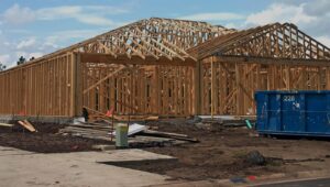 A blue dumpster filled with construction debris next to a house in the framing stage, provided by Discount Dumpster in Naples, FL.