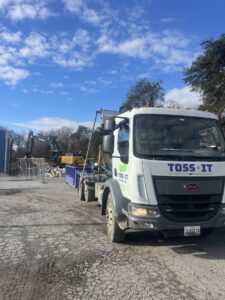 A Toss-It truck with a dumpster at a construction site with an excavator in the background in Glen Burnie, MD.