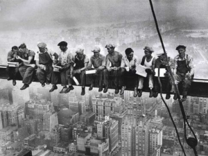 Construction workers having lunch on a steel beam high above a city, representing the work of Behar Construction in Miami, FL