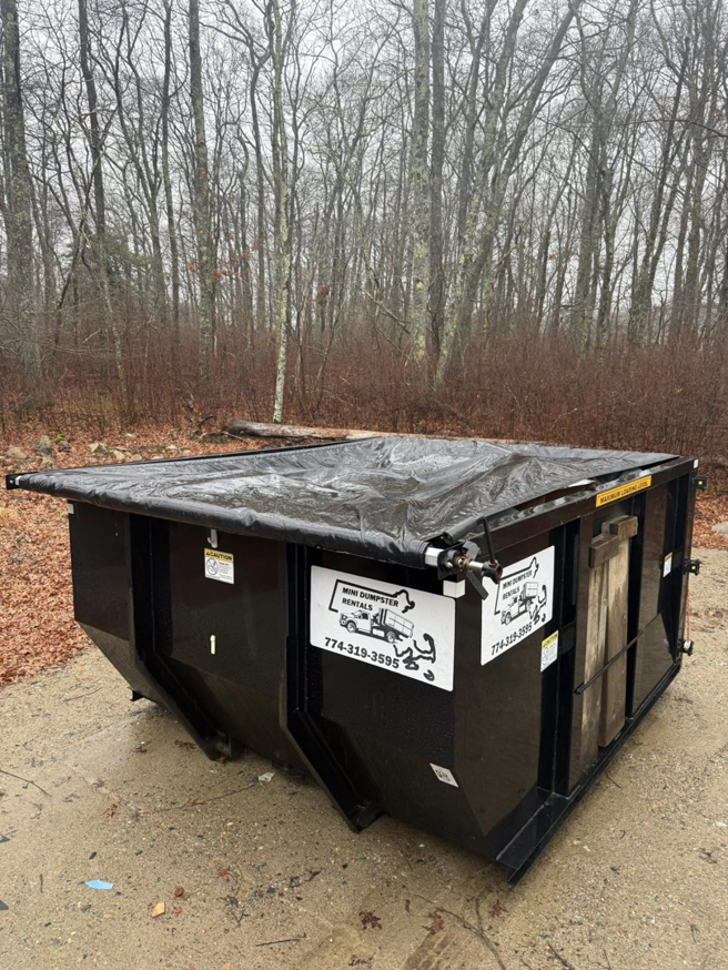 A black dumpster with a tarp cover from Mini Dumpster Rentals, situated in a forest setting in New Bedford, MA.