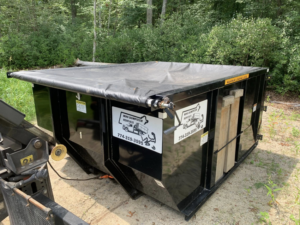 A black dumpster with a protective tarp cover from Mini Dumpster Rentals, located in a wooded area in New Bedford, MA.