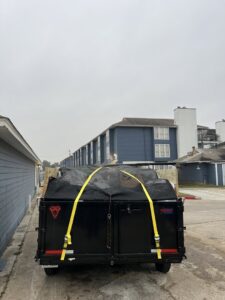 The back of a dump trailer, covered and secured with straps, filled with junk outside an apartment complex by Tidy Loaders Junk Removal in Houston, TX.