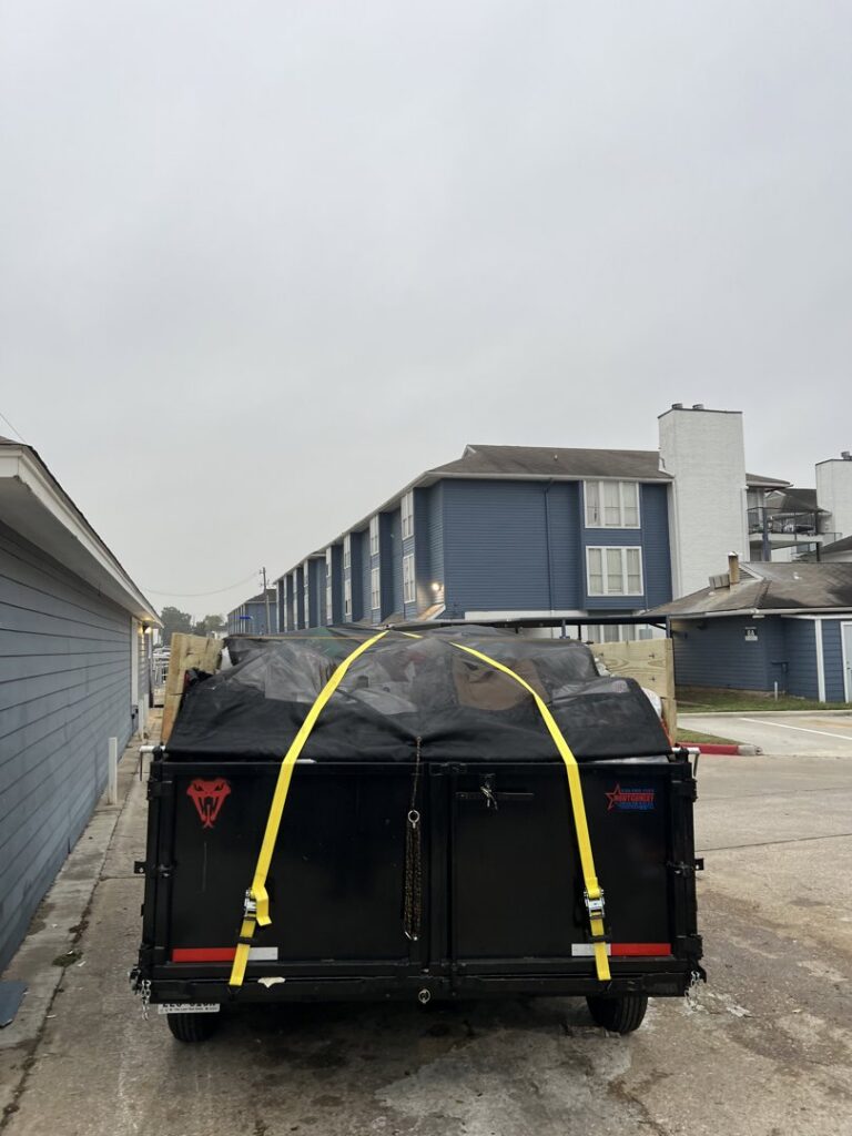 The back of a dump trailer, covered and secured with straps, filled with junk outside an apartment complex by Tidy Loaders Junk Removal in Houston, TX.