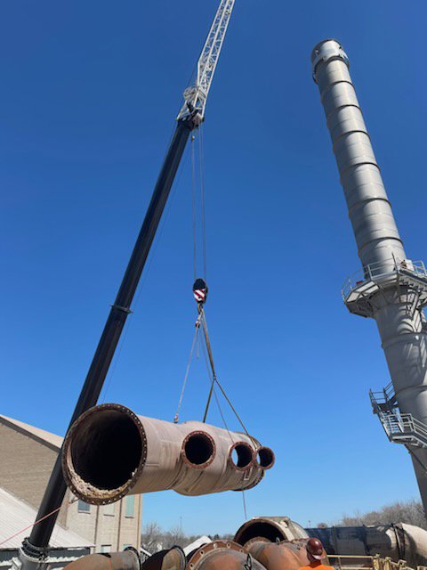 A crane lifting large industrial pipes next to a tall structure for Midwest Crane & Rigging Inc. in Belvidere, IL