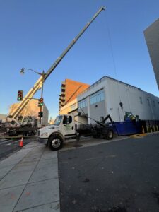 A crane loading construction debris into a Toss-It dumpster on a city street in Glen Burnie, MD.