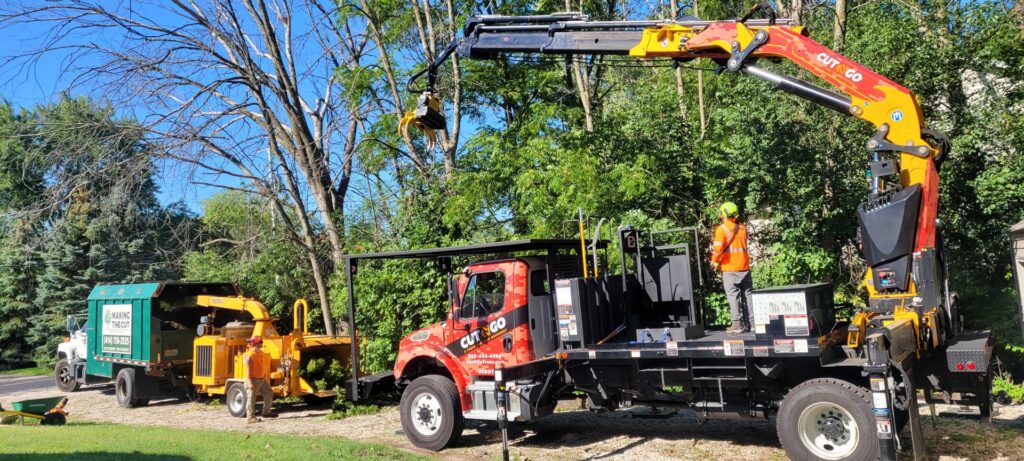 A crane truck with a grapple, chipper, and chipper truck on a tree service job site by Making the Cut Tree Service in Muskego, WI.