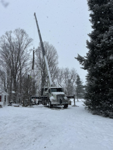 A crane truck lifting a large tree trunk section during removal in a snowy residential area by DJ's Tree Service in Colchester, VT.
