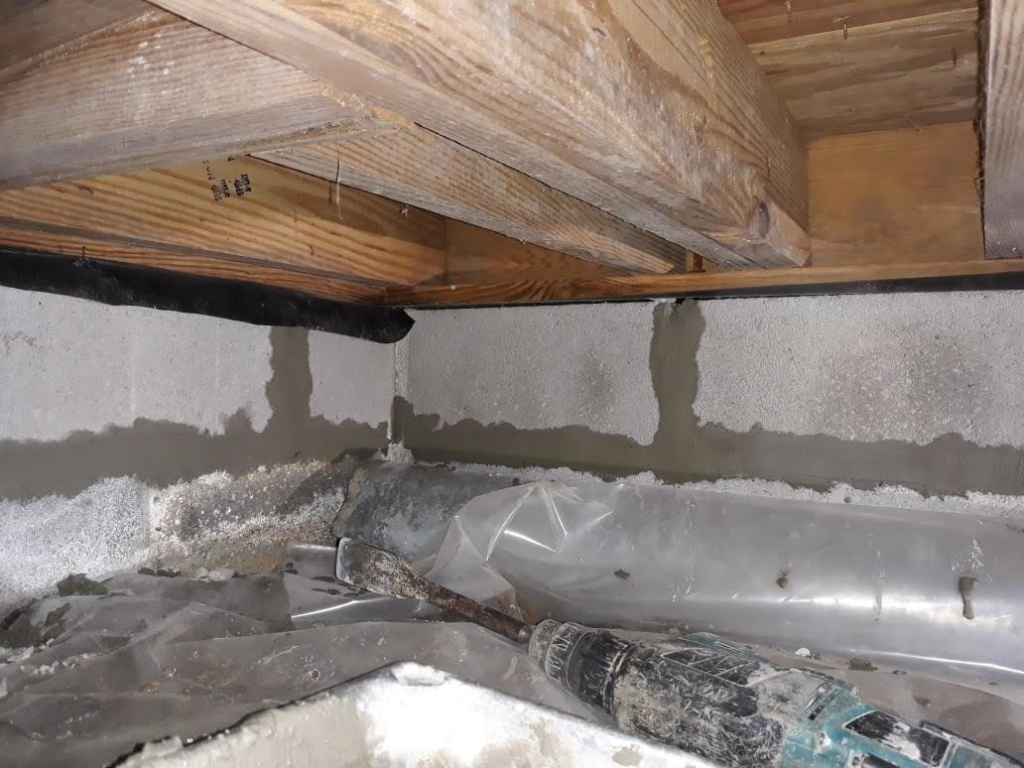 Crawl space foundation repair showing wooden joists, concrete walls, and a drill with fresh sealant by Ram Jack Foundation Solutions in Hampton, VA.