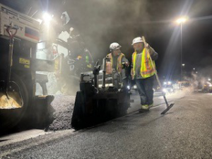 A crew operating an asphalt paver to lay new pavement at night for Northeast-Paving in Bangor, ME