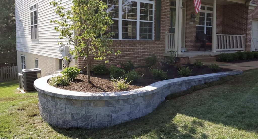 A curved stone retaining wall with landscaping around a tree in front of a house by Mincho Contractor LLC in Jersey City, NJ